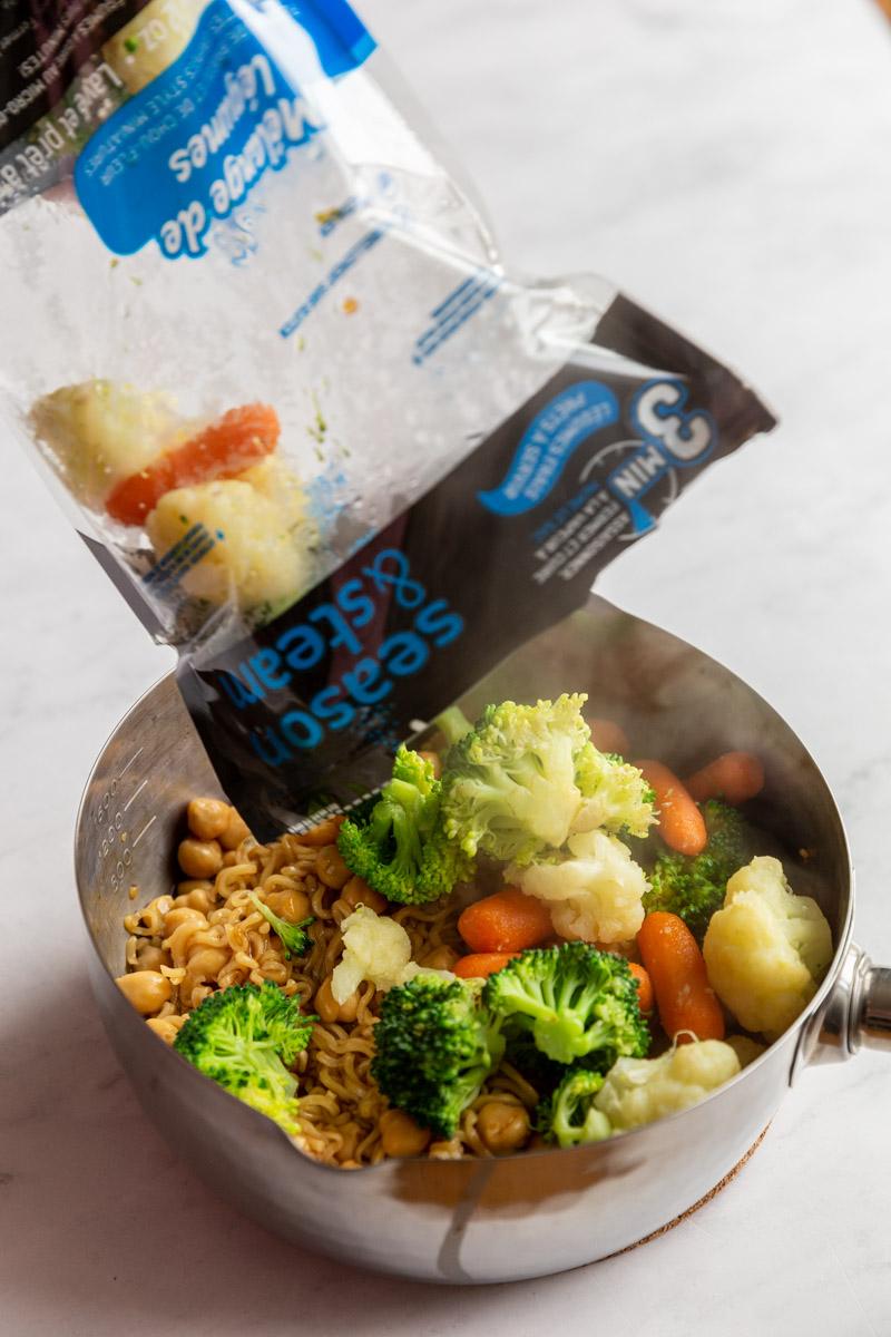 bag of ocean mist season and steam veggies being poured in a saucepan with cooked ramen noodles