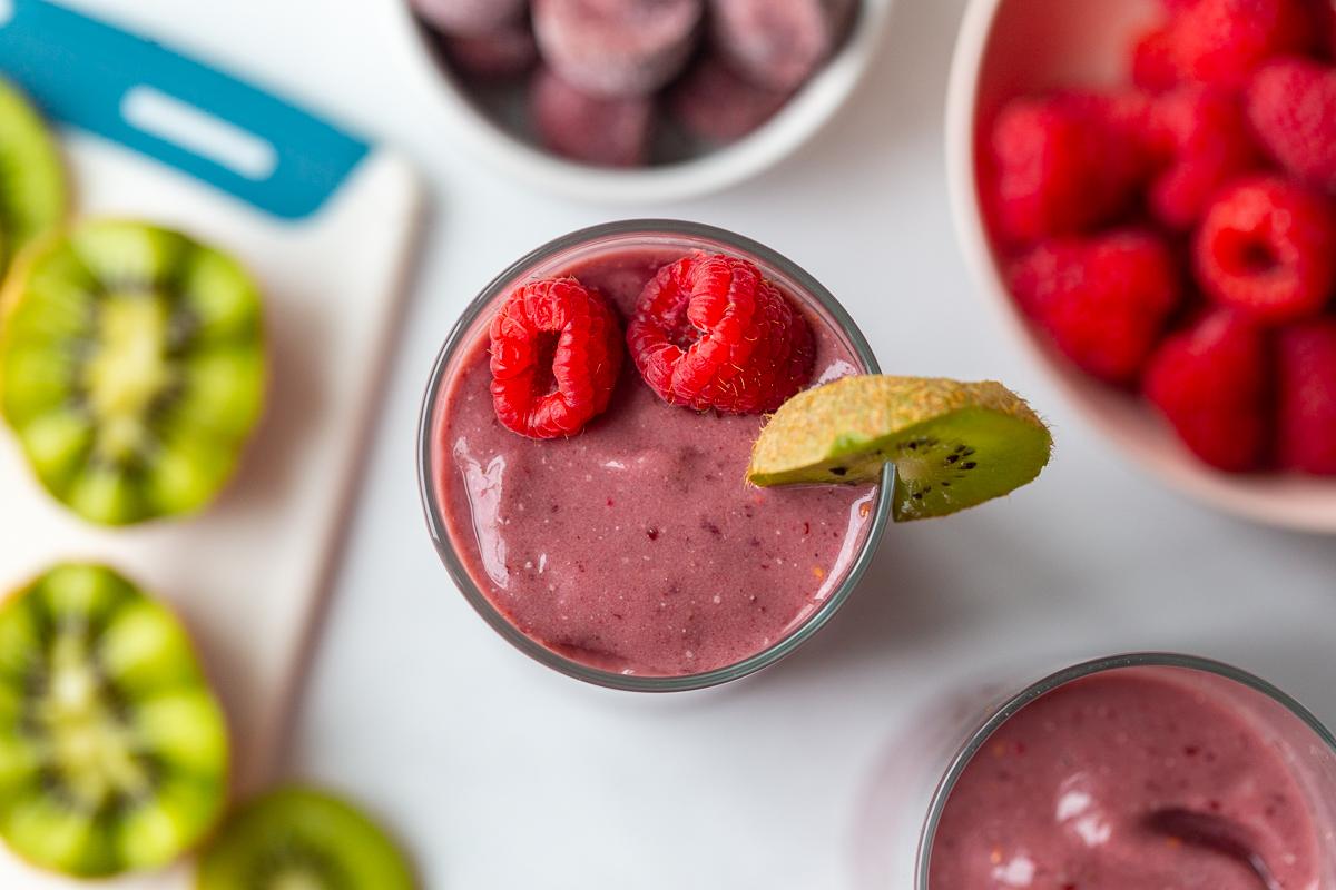 acai smoothie in a tall glass topped with raspberry and kiwi with more kiwi in the background