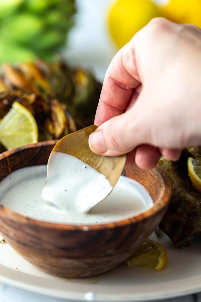 piece of artichoke being dipped in a white sauce with full artichokes in the background