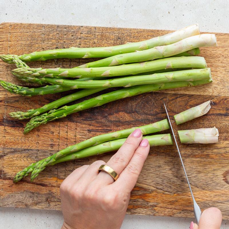 asparagus on a wood cutting board with a hand cutting off the woody ends of the asparagus
