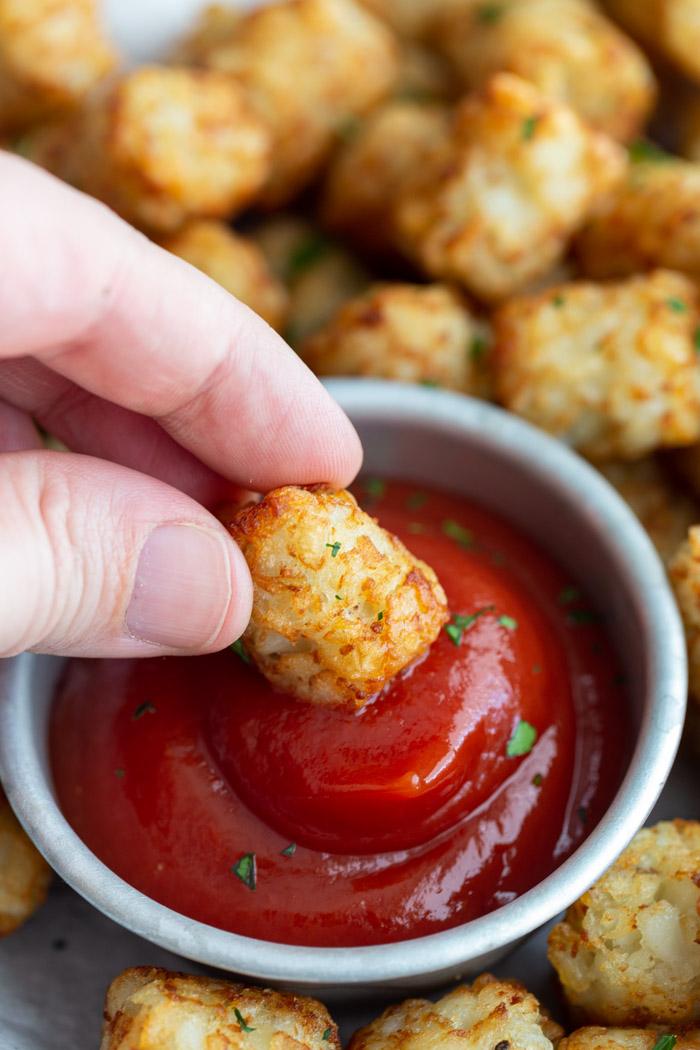 hand dipping a tater tot into a small bowl of ketchup with more tater tots in the background