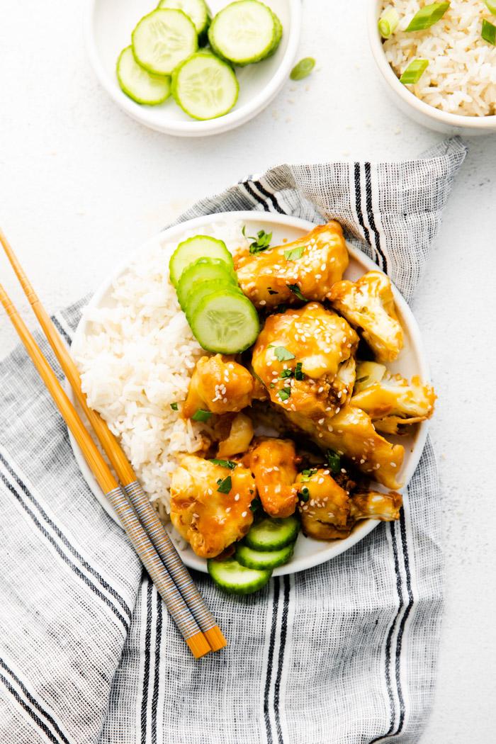 plate filled with white rice, cucumber, and sticky sesame cauliflower. plate on top of a towel with chopsticks on the side