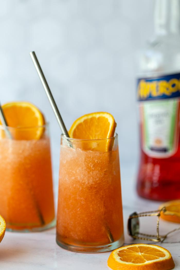 2 frozen aperol spritz cocktails with orange slices in them and a bottle of aperol in the background