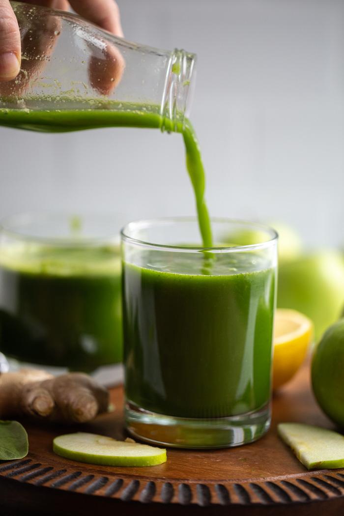 Apple green juice being poured into a small glass on a wood board