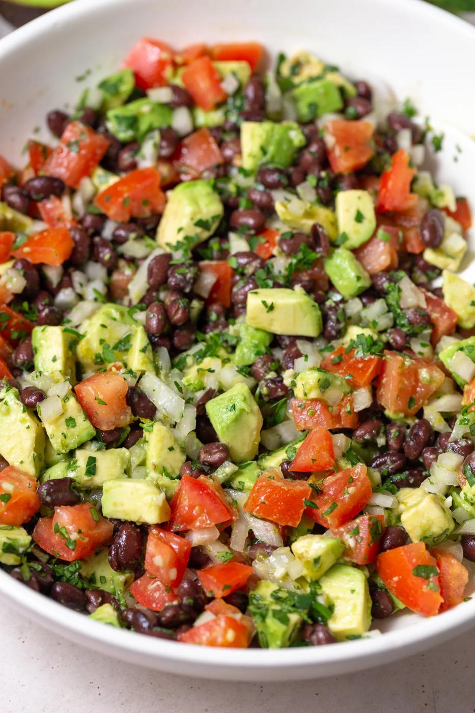large white bowl filled with an Avocado & Black Bean Salad that's sprinkled with cilantro. Some avocados and cilantro in the corn of the shot