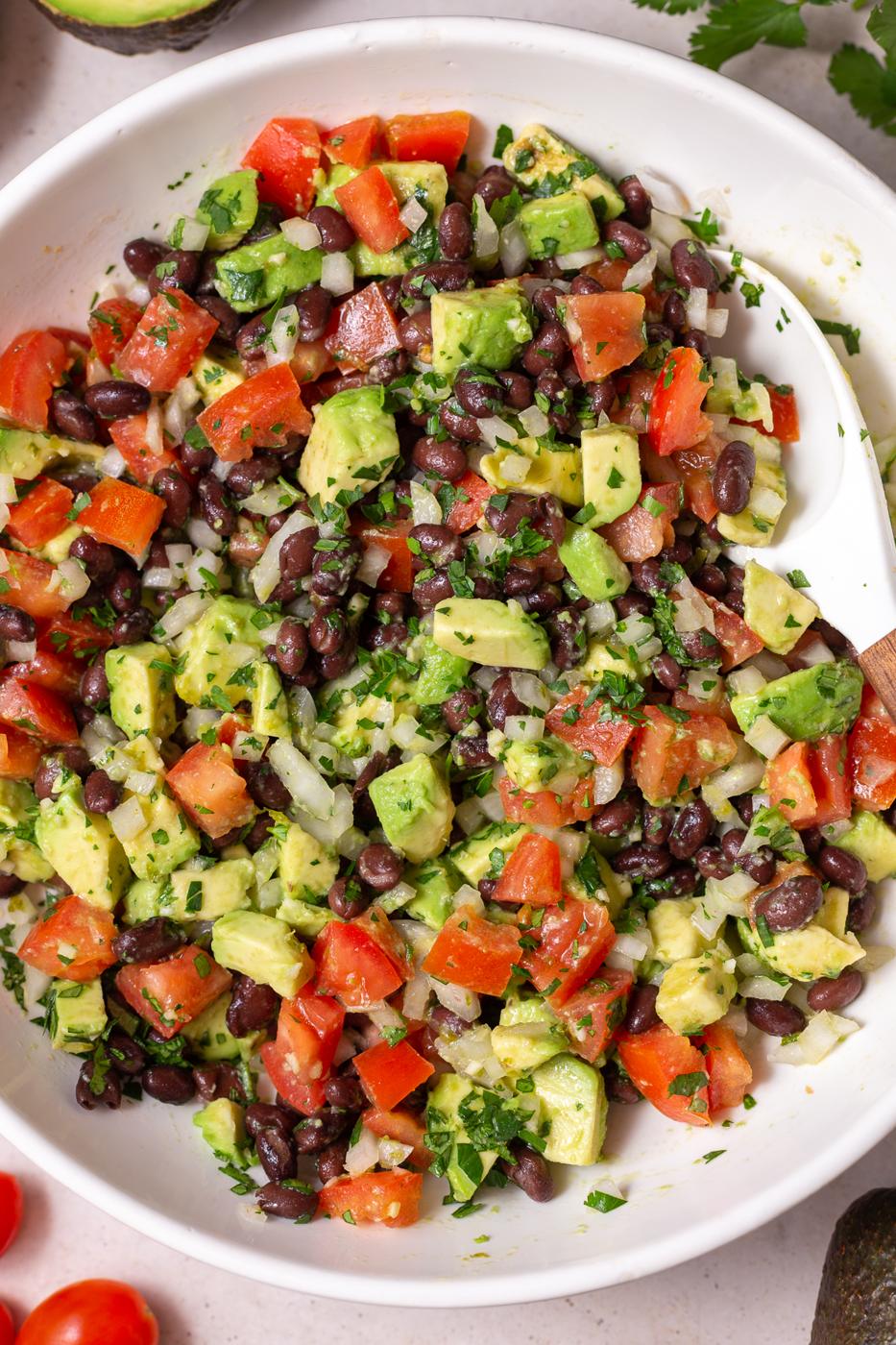 large white bowl filled with an Avocado & Black Bean Salad that's sprinkled with cilantro. Some avocados and cilantro in the corn of the shot