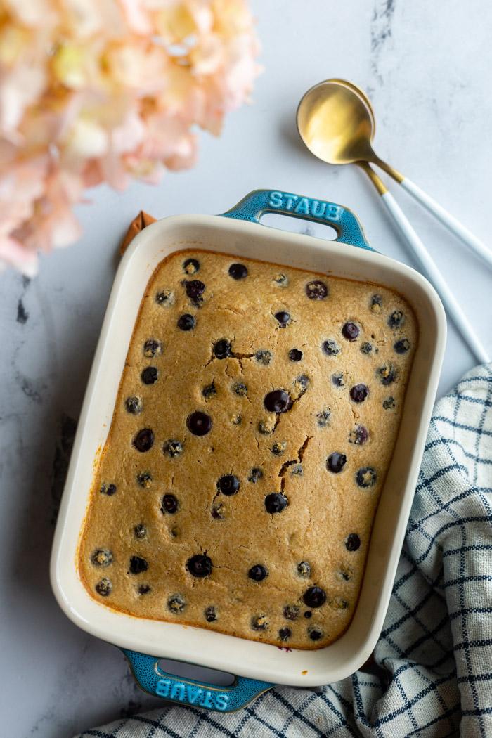 baked oats with blueberries in a small casserole dish with a napkin and spoons on the side. pink flowers in the corner of the shot