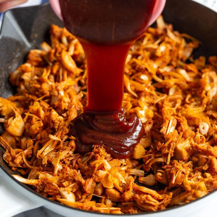 bbq sauce being poured on shredded jackfruit in a skillet