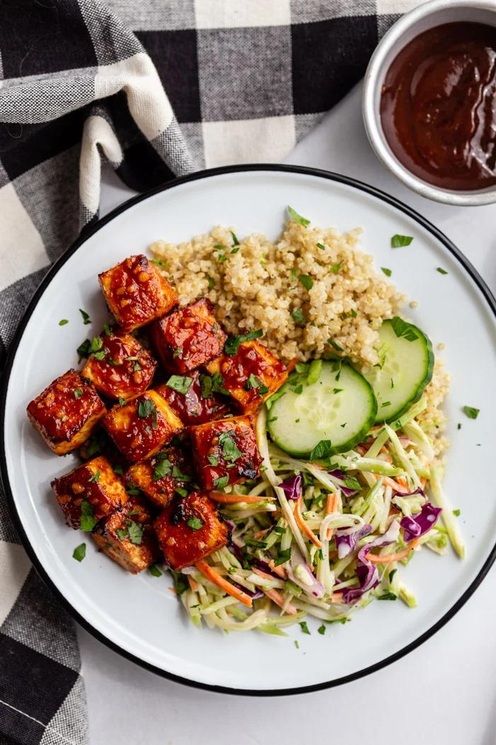 white bowl with quinoa, coleslaw, and bbq tofu in it plus a few pieces of cucumber