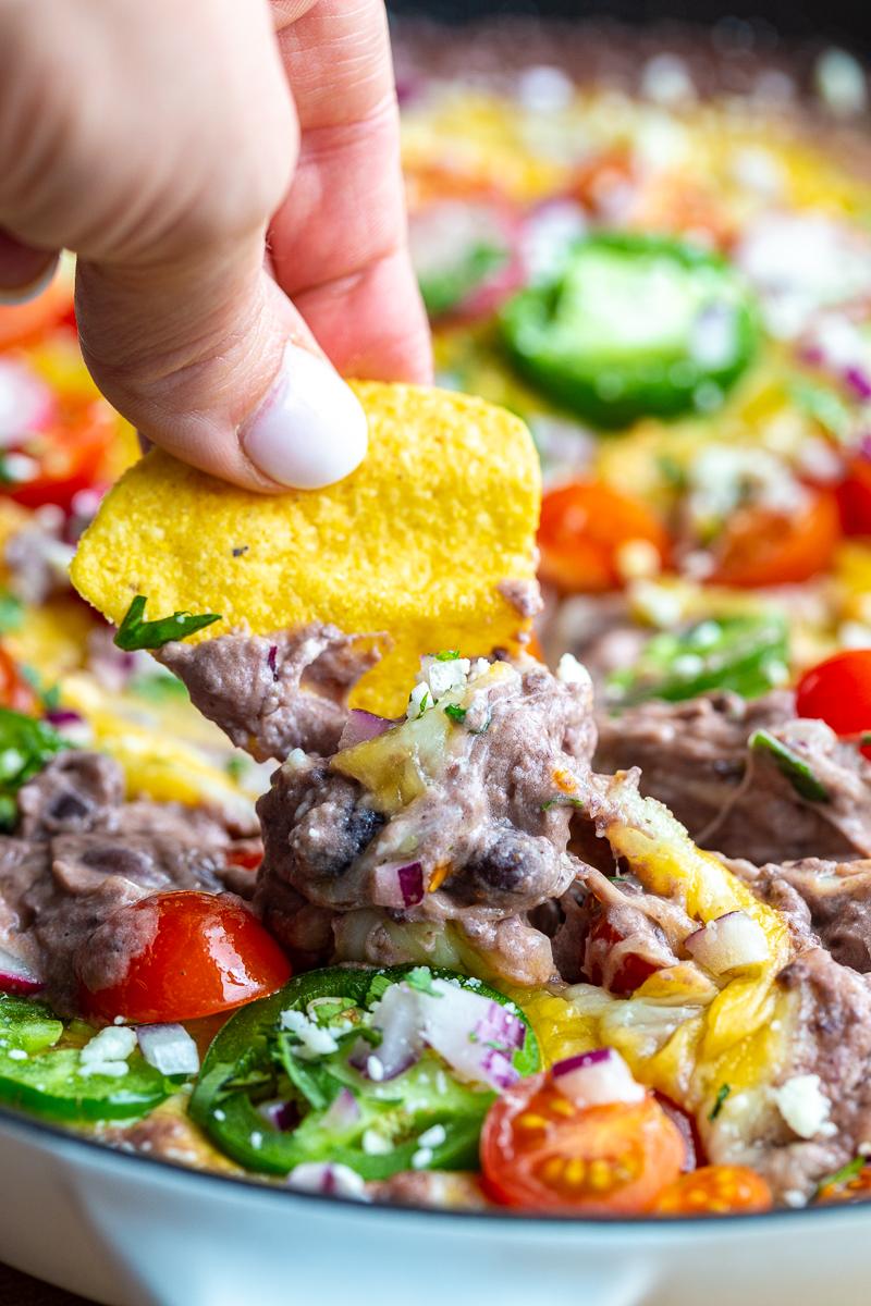 black bean dip in a cast iron. It's covered with melted cheese and topped with cherry tomatoes, sliced jalapeno, diced red onion, cotija cheese, sliced radishes, and chopped cilantro. There's a hand holding a tortilla chip up that's been dipped in the dip