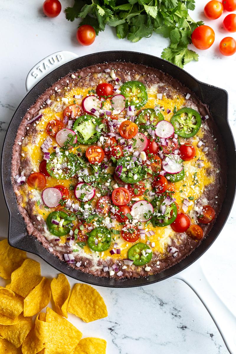 black bean dip in a cast iron. It's covered with melted cheese and topped with cherry tomatoes, sliced jalapeno, diced red onion, cotija cheese, sliced radishes, and chopped cilantro
