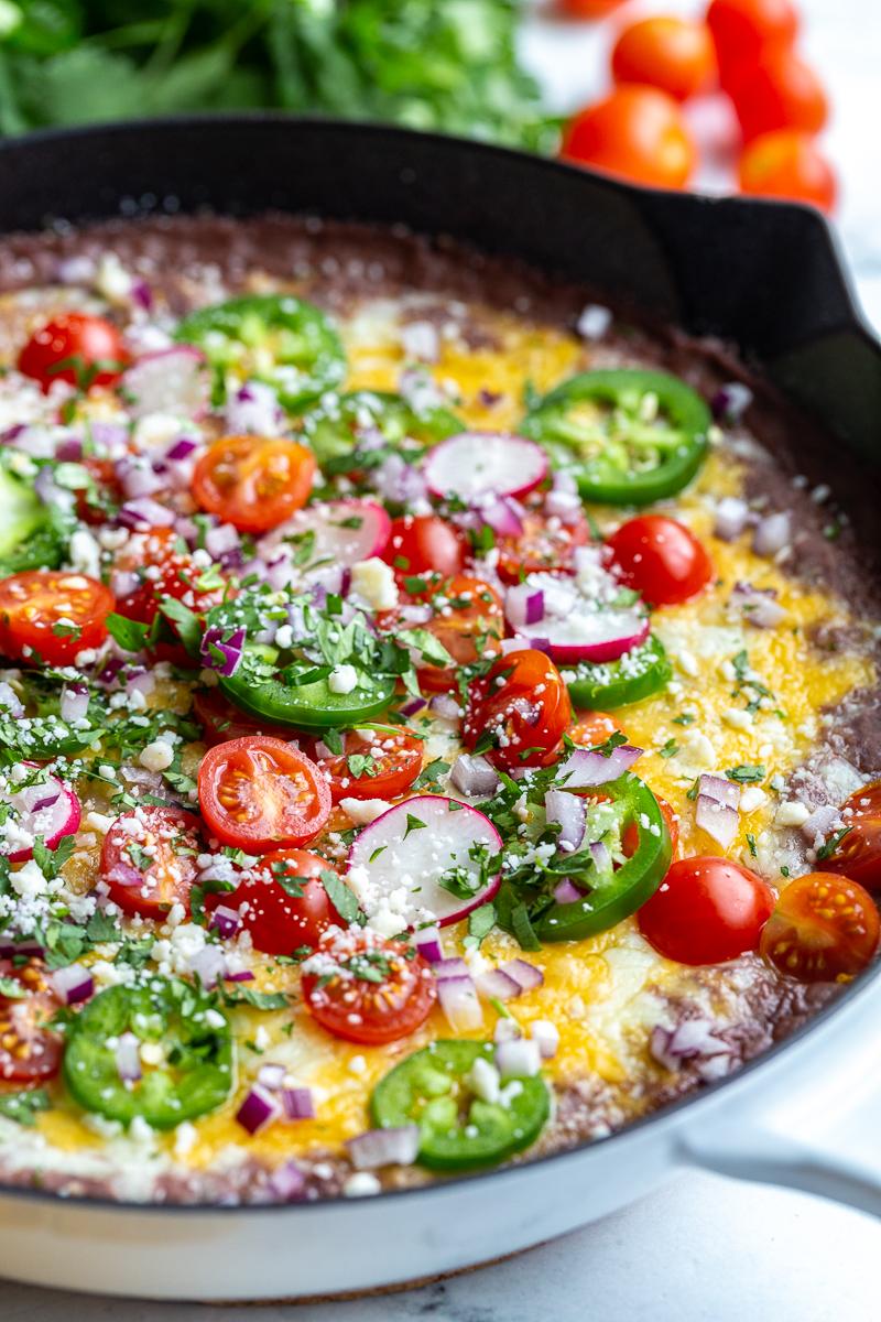 side shot of black bean dip in a cast iron. It's covered with melted cheese and topped with cherry tomatoes, sliced jalapeno, diced red onion, cotija cheese, sliced radishes, and chopped cilantro