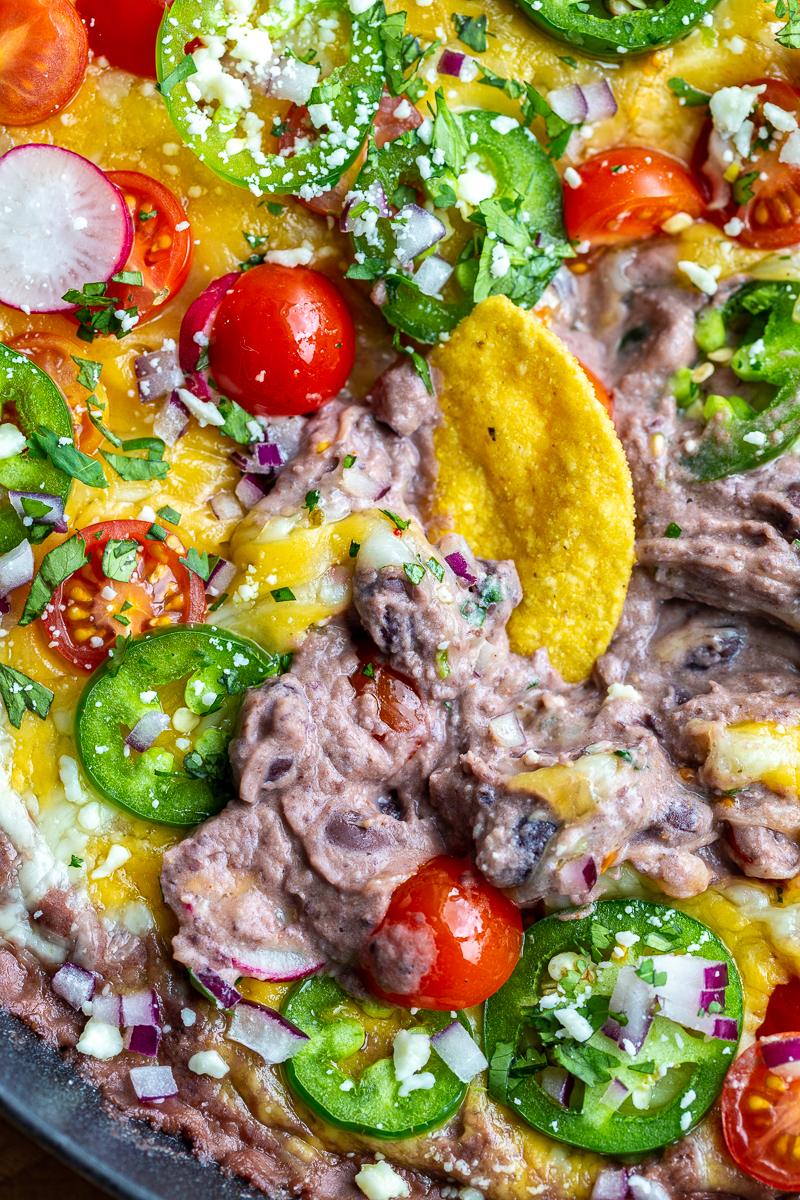 slow up shot of black bean dip in a cast iron. It's covered with melted cheese and topped with cherry tomatoes, sliced jalapeno, diced red onion, cotija cheese, sliced radishes, and chopped cilantro. There's a tortilla chip sitting in the dip