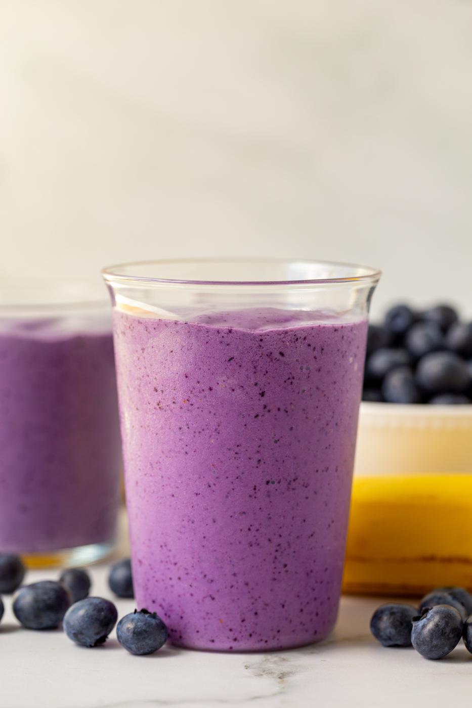 blueberry smoothie in a medium clear glass with blueberries and bananas in the background