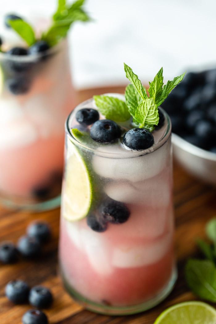 side shot of 2 blueberry mojitos in clear glass with ice and fresh blueberries, mint, and lime garnishing the glass. Both sitting on a wood board with ingredients around