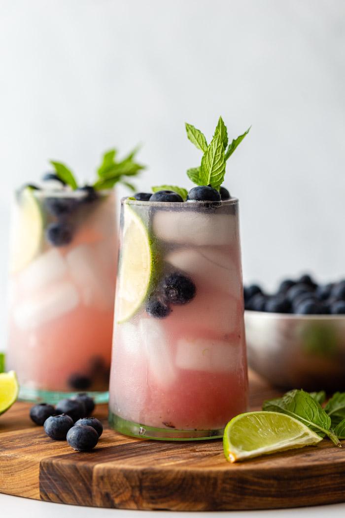 side shot of 2 blueberry mojitos in clear glass with ice and fresh blueberries, mint, and lime garnishing the glass. Both sitting on a wood board with ingredients around