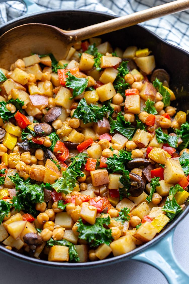 using a wooden spoon to stir potatoes and veggies in a large skillet