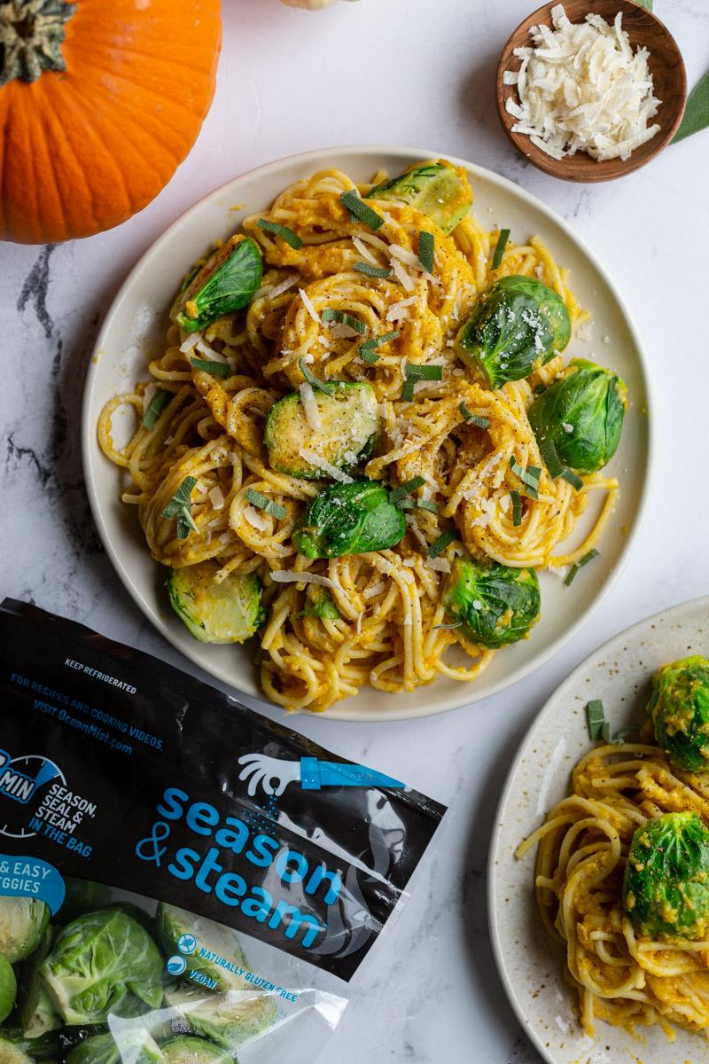 creamy pumpkin pasta spaghetti on a plate with brussels on top and small pumpkins in the corner of the shot and a bag of ocean mist sprouts in the corner