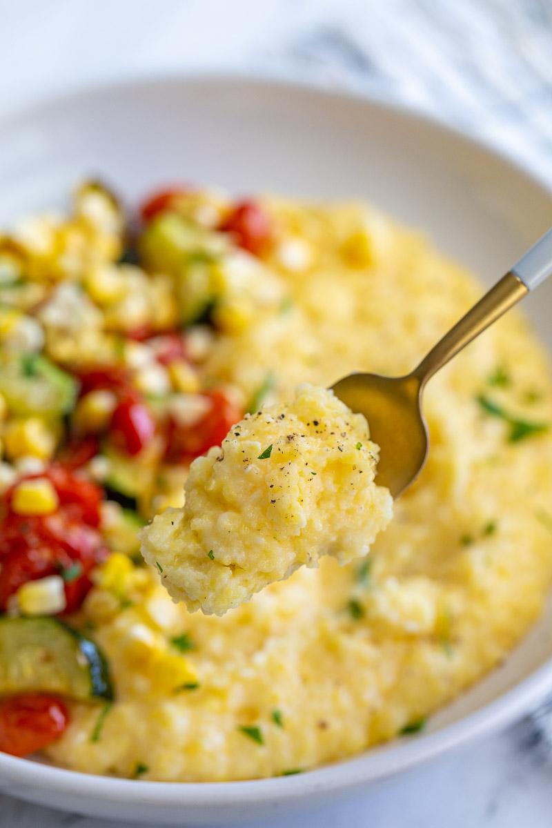 bowl of cheesy polenta topped with roasted zucchini, corn, and tomatoes and sprinkled with fresh herbs with a fork holding up a bite of the polenta