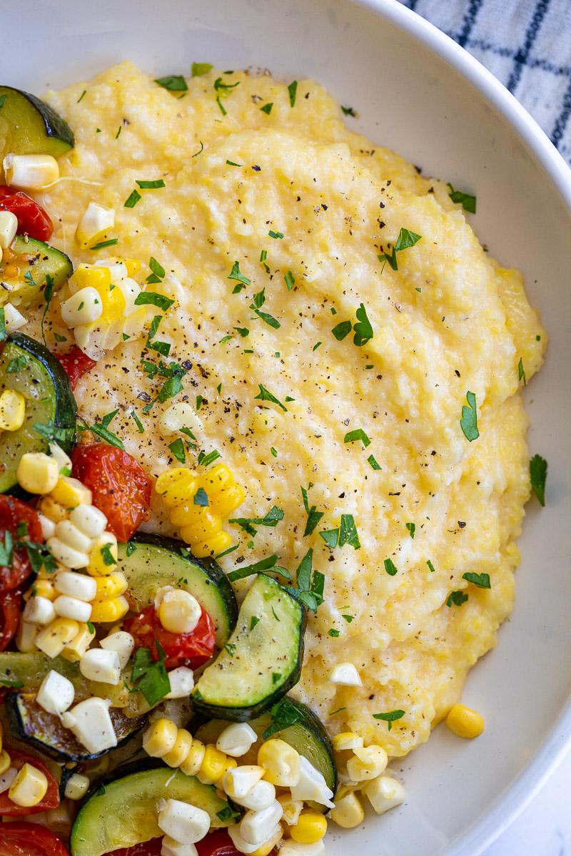 up close shot of a bowl of cheesy polenta topped with roasted zucchini, corn, and tomatoes and sprinkled with fresh herbs