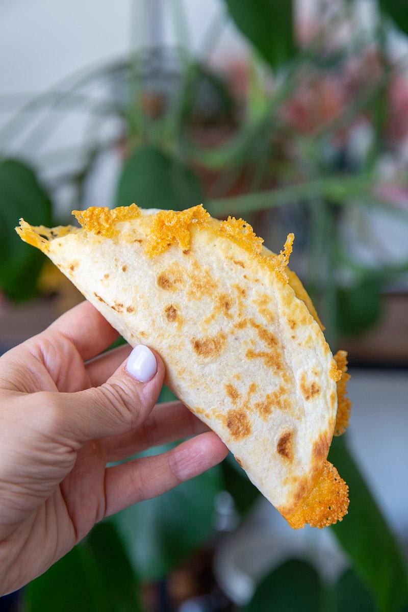 hand holding up a taco shell that's lightly browned with crispy cheese all around the edges