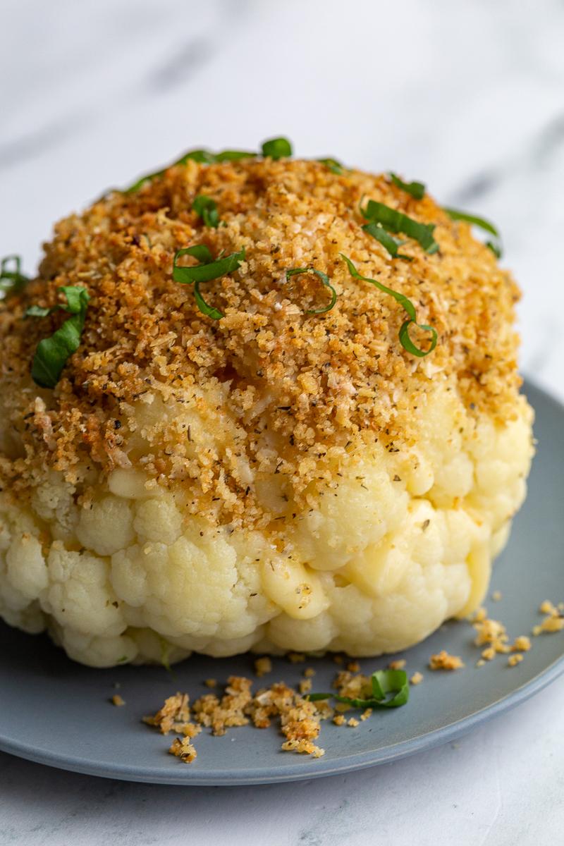 whole head of cauliflower sitting on blue dish. It has white cheesy oozing out the sides and it's topped with a toasted breadcrumb mixture and fresh basil ribbons