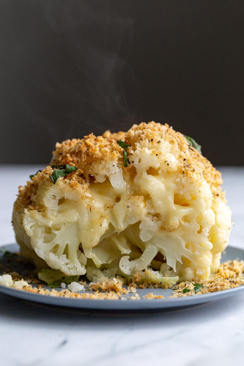 whole head of cauliflower sitting in blue dish. It's been cut in half and has white cheesy oozing out the middle and it's topped with a toasted breadcrumb mixture and fresh basil ribbons