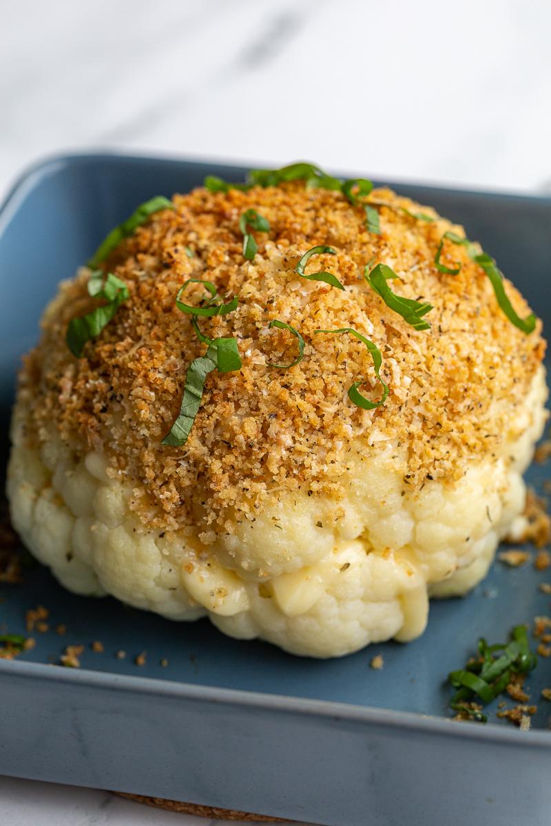 whole head of cauliflower sitting in blue baking dish. It has white cheesy oozing out the sides and it's topped with a toasted breadcrumb mixture and fresh basil ribbons