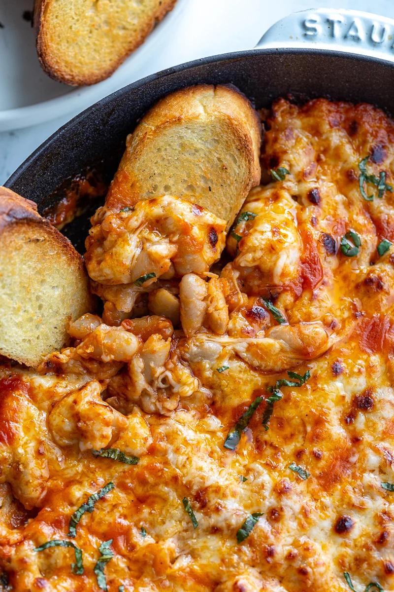 white bean tomato bake topped with lots of white cheese and fresh basil ribbons. In a white cast iron skillet with toasted bread on the side and some of the beans scooped onto the bread