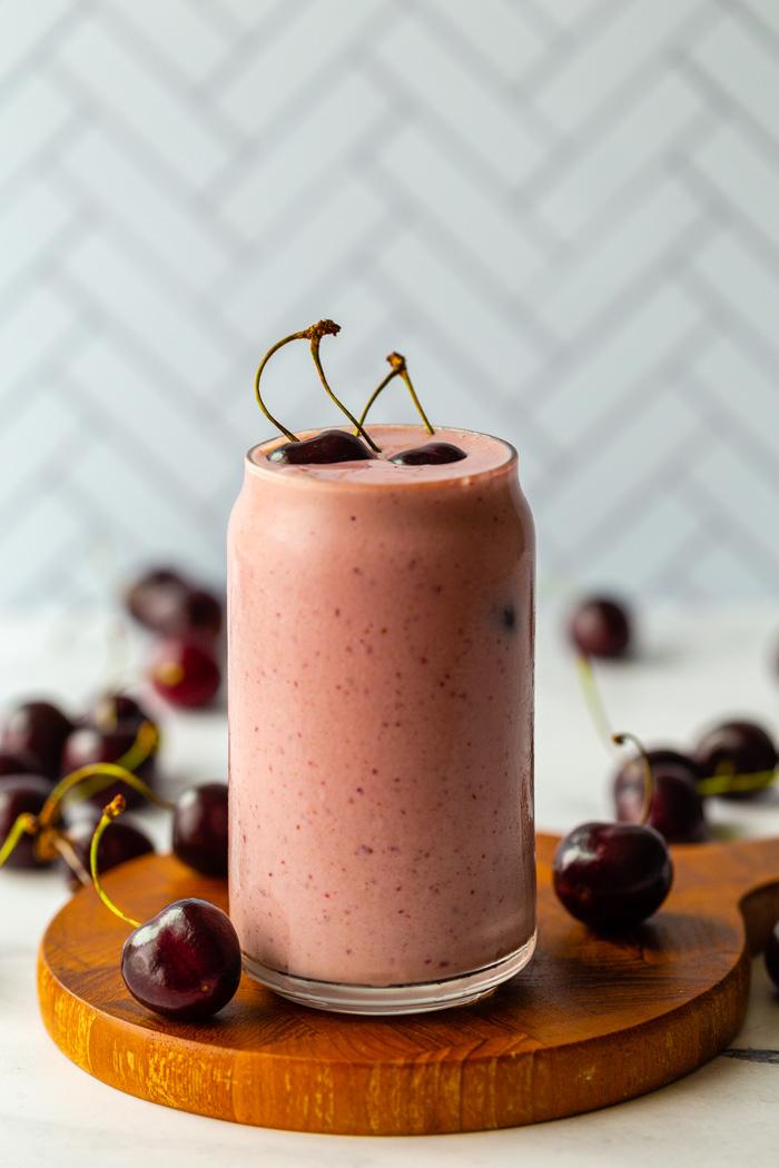 clear glass filled with a pink cherry smoothie with fresh cherries on top sitting on a wood board. tile surface in the background