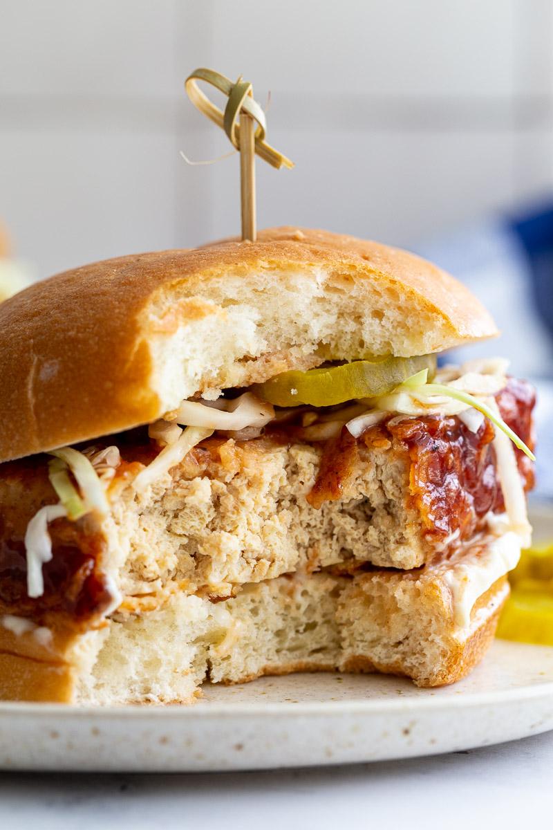 tofu "chicken sandwich on a bun with mayo, cabbage and pickles and a toothpick in the center. A bite is taken out so you can see the center