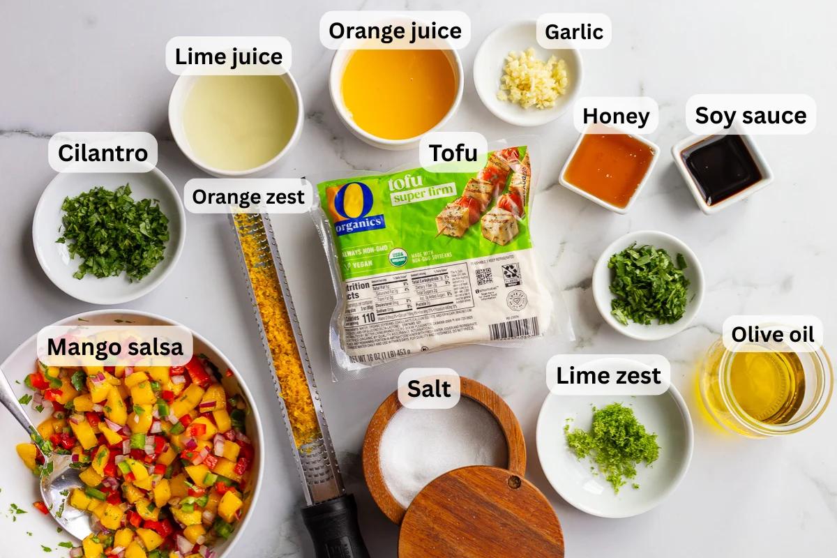 Overhead shot of cilantro lime tofu ingredients including especially firm tofu, lime juice, lime zest, orange juice, orange zest, garlic, honey, soy sauce, cilantro, olive oil, salt, and a bowl of mango salsa, each labeled on a marble countertop.