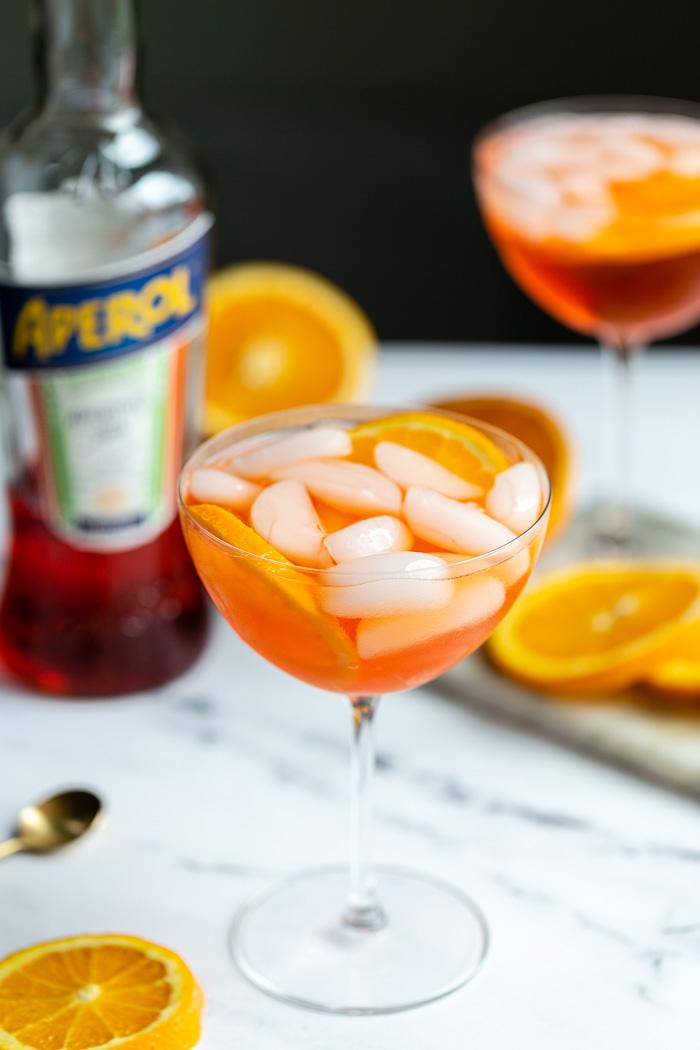 orange cocktail in a wine glass on a marble backdrop with oranges all around and a bottle of aperol in the background