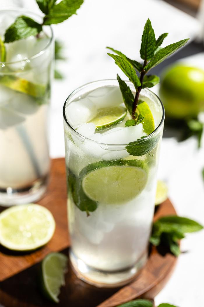 2 mojitos in tall glasses with fresh limes and mint in them. Sitting on a round wood board
