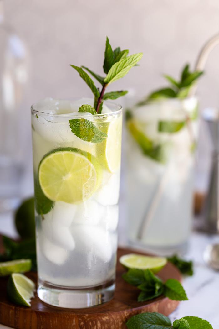 2 mojitos in tall glasses with fresh limes and mint in them. Sitting on a round wood board with a light tile background