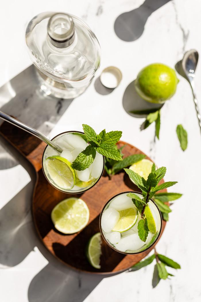 top down shot 2 mojitos in tall glasses with fresh limes and mint in them. Sitting on a round wood board