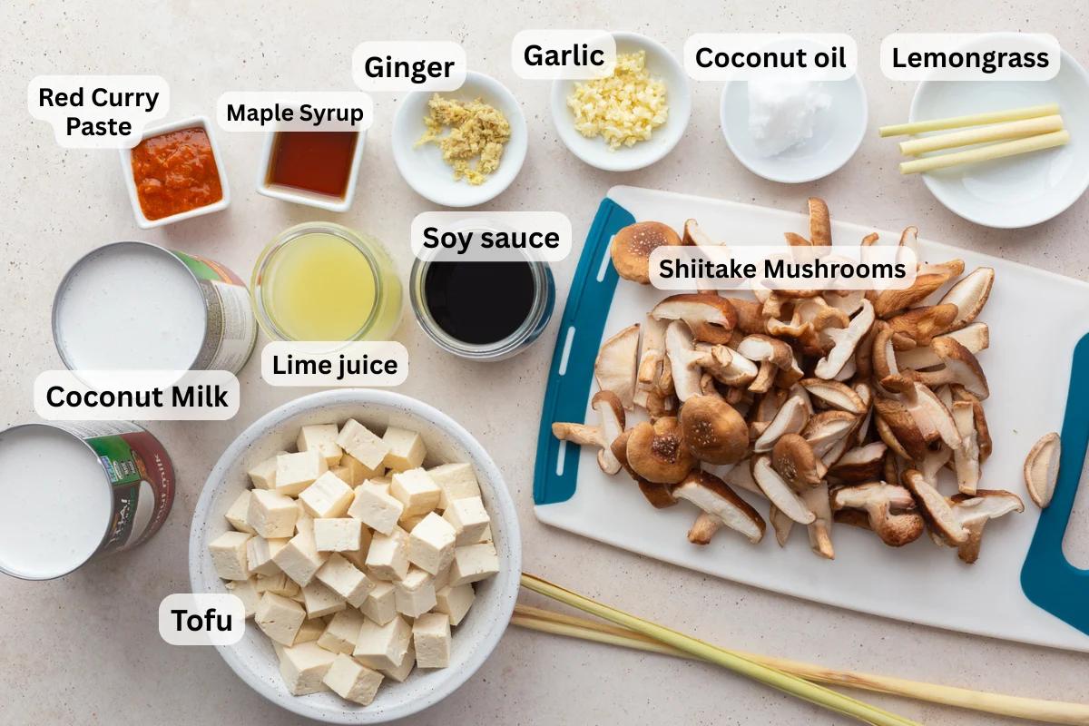 labeled ingredients needed to make coconut tofu soup including coconut milk, lime juice, shiitake mushrooms, red curry paste, tofu, ginger, garlic, lemongrass, and soy sauce