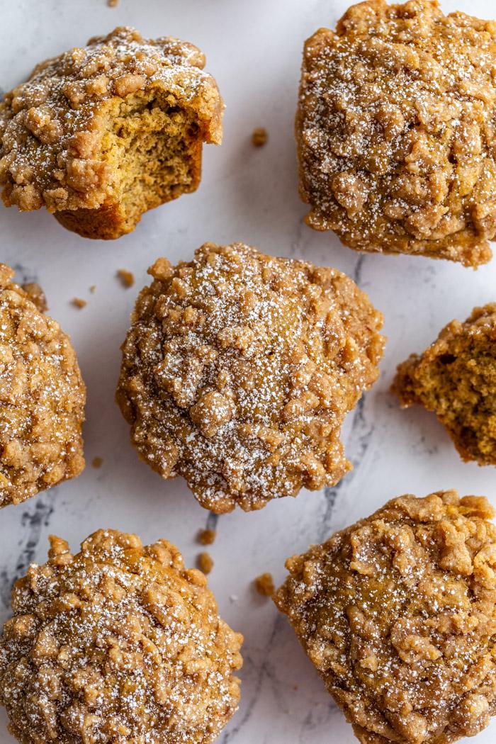 top down shot of banana muffins with crumble topping and a dusting of powdered sugar on top. More muffins all around laying on a marble board