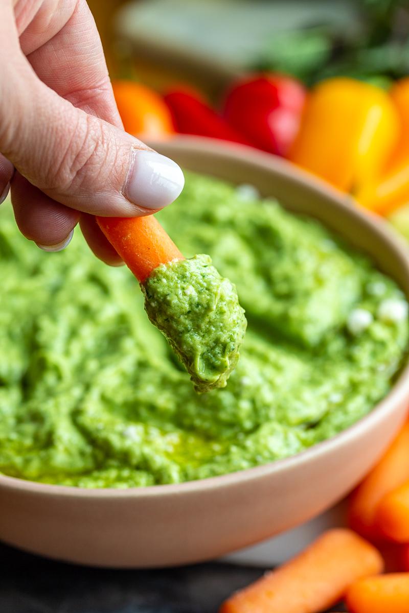 Green goddess dip (a bright green herby dip) in a pink bowl. This shot has a hand holding a baby carrot that has some of the dip on it