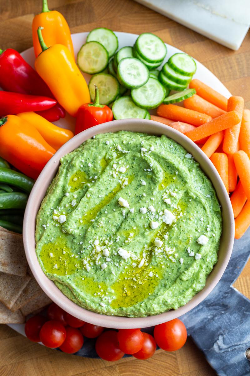 Green goddess dip (a bright green herby dip) in a pink bowl. there's feta sprinkled on top and lots of fresh veggies around the bowl