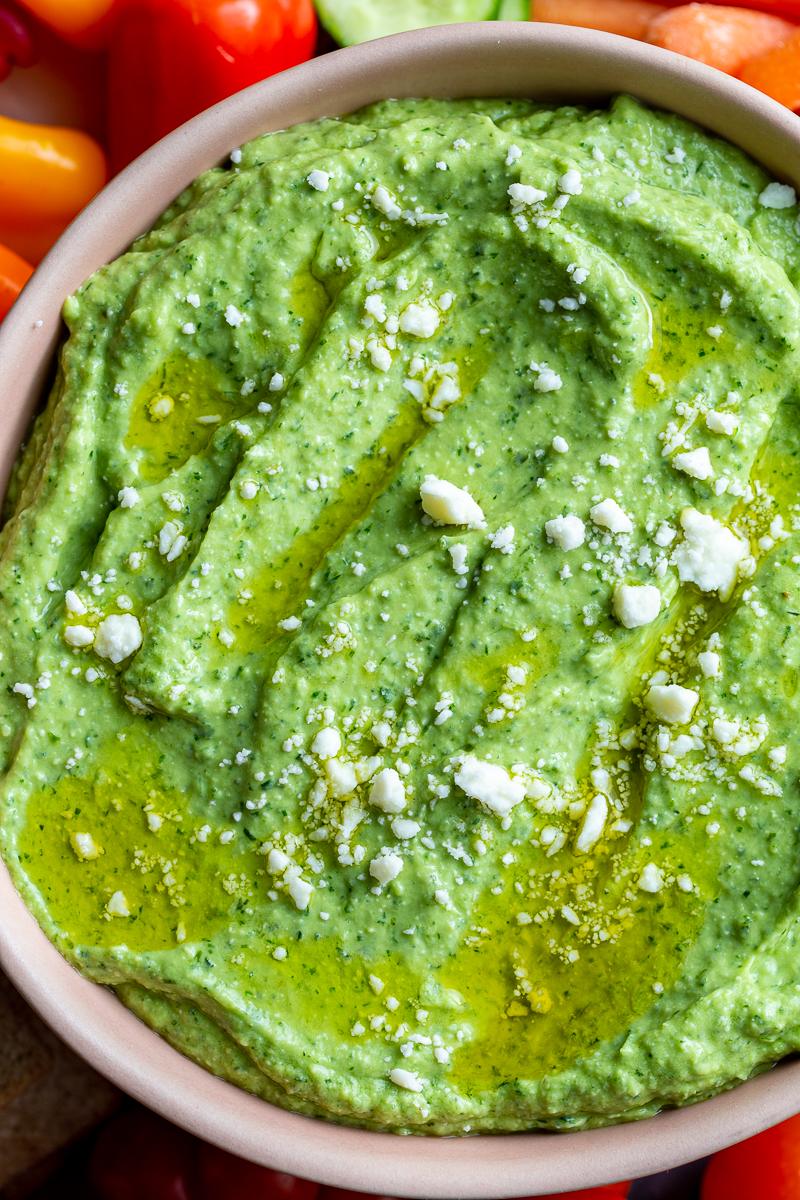 Green goddess dip (a bright green herby dip) in a pink bowl. there's feta sprinkled on top and lots of fresh veggies around the bowl
