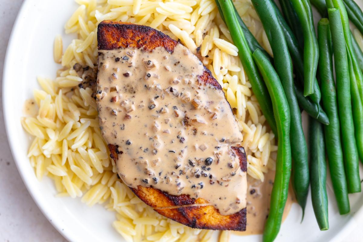 tofu "steak" laying on a bed of orzo, next to green beans, and covered in a creamy peppercorn sauce