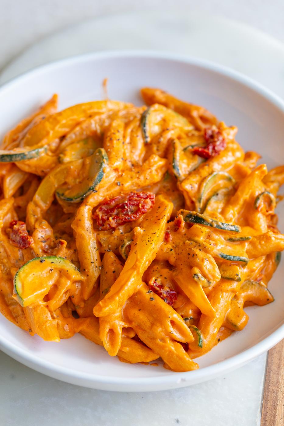 Creamy tomato pasta with bell peppers and zucchini in a white bowl.