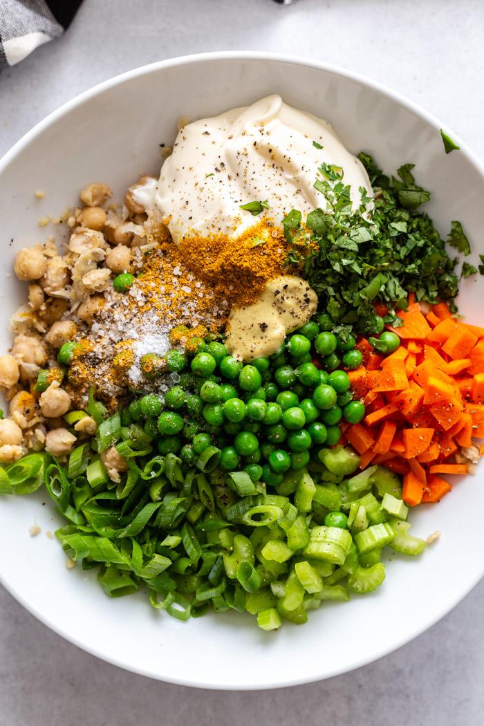 large white bowl filled with the ingredients for chickpea salad including chickpeas, green onion, peas, carrots, celery, mayo, cilantro, dijon, and curry powder