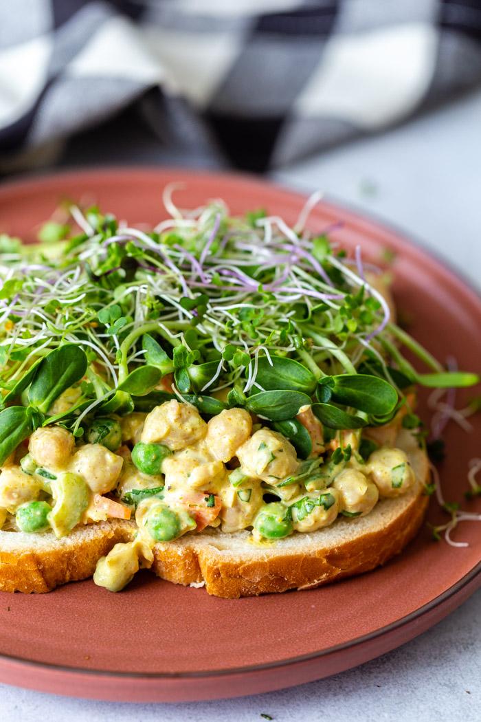 red plate with a curried chickpea salad sandwich on top that's topped with microgreens