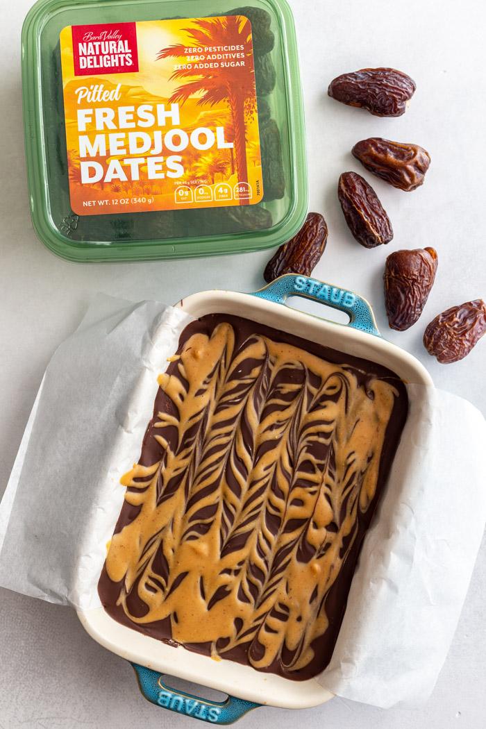 pan filled with date bars that are covered in chocolate and peanut butter drizzle. a green carton of natural delights medjool dates in the frame