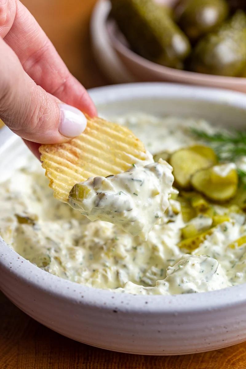 Dill pickle dip in a bowl topped with bright green diced pickles. There's a hand hold a chip up that has some of the dip on it