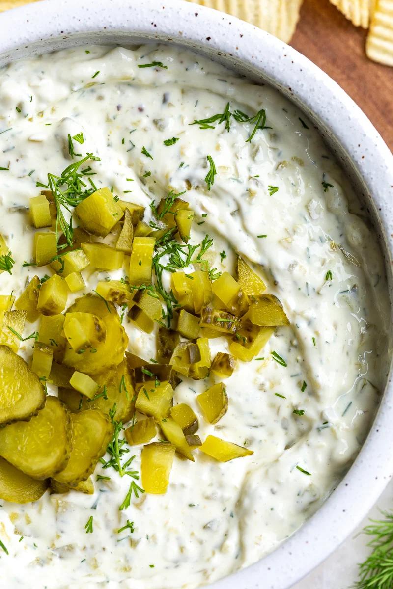 Dill pickle dip in a bowl topped with bright green diced pickles. There are ruffles chips in the corner of the shot