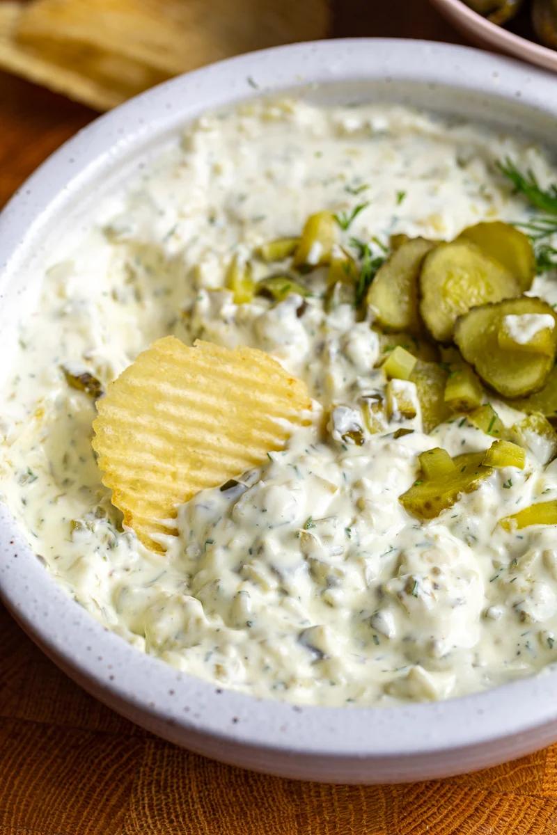 Dill pickle dip in a bowl topped with bright green diced pickles. There is a chip dipped in the bowl