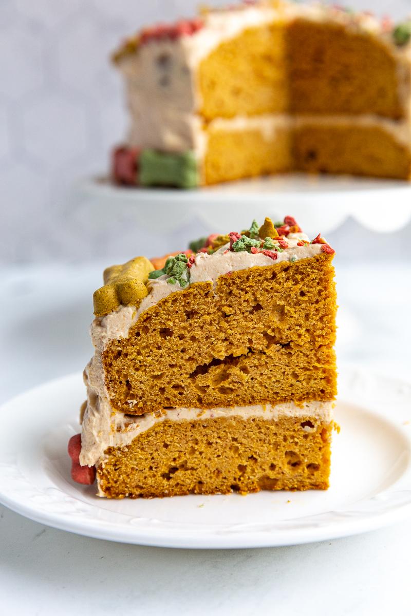 slice of dog birthday cake on a plate with the rest of the cake on a cake stand in the background. the cake has colorful bones on iti